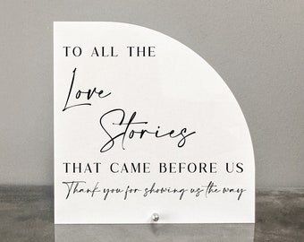 May include: White acrylic sign with black text that reads "To all the Love Stories That Came Before Us Thank you for showing us the way".