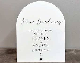 Op de afbeelding: Een wit, boogvormig bord met de tekst "to our loved ones" in zwarte cursieve letters. Het bord bevat ook de tekst "WHO ARE DANCING WITH US IN HEAVEN we love AND MISS YOU" in zwarte letters met een hartje onderaan.