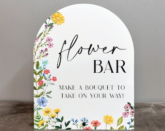 May include: A white sign with a floral border and the text "flower bar" in black script. Below the text is the phrase "Make a bouquet to take on your way!" in black.