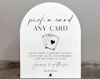 Könnte beinhalten: Ein weißes Schild mit abgerundeter Oberseite, auf dem "pick a card ANY CARD" steht, mit einer schwarzen Illustration von Spielkarten. Unter dem Text steht eine Nachricht, die lautet: "PLEASE WRITE A MESSAGE AND SIGN YOUR NAME, SO WE CAN REMEMBER THIS DAY WHEN WE PLAY!" mit den Namen "James & Allison" und dem Datum "09.27.2024".