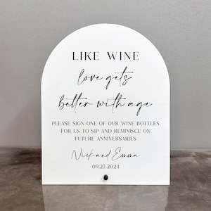 May include: White arched sign with black text that reads "LIKE WINE love gets better with age." The sign also includes the names "Nick and Emma" and the date "09.27.2024."