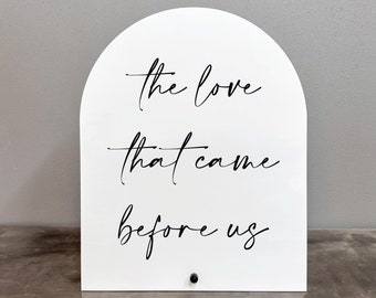 May include: White arch-shaped sign with black calligraphy that reads "the love that came before us".
