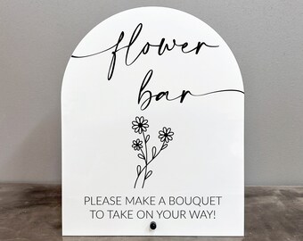 May include: White sign with black text that reads "flower bar" with a small flower illustration. The sign also says "Please make a bouquet to take on your way!"