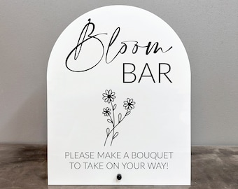 May include: White sign with an arched top that says "Bloom Bar" in black cursive lettering. Below the text is a black line drawing of a bouquet of flowers. The sign also says "Please make a bouquet to take on your way!"