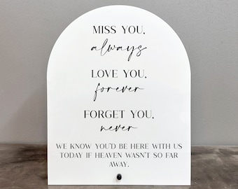 Op de afbeelding: Een wit boogvormig bord met zwarte tekst die luidt: "Miss you, always. Love you, forever. Forget you, never. We know you'd be here with us today if heaven wasn't so far away."