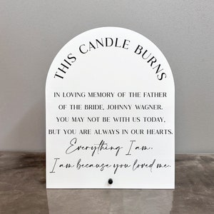 May include: White acrylic sign with a rounded top that reads "This candle burns in loving memory of the father of the bride, Johnny Wagner. You may not be with us today, but you are always in our hearts. Everything I am. I am because you loved me."