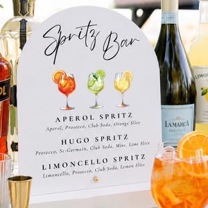 May include: A white arched sign labeled Spritz Bar with illustrations of three cocktails, listing ingredients for Aperol, Hugo, and Limoncello Spritz. Bottles of Aperol, Prosecco, and Limoncello are displayed alongside three filled cocktail glasses.
