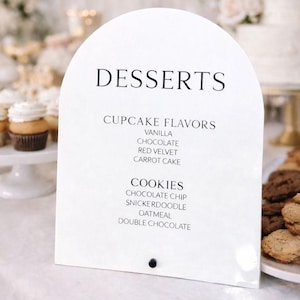 May include: A white, arched sign with the word "DESSERTS" at the top. Below, it lists cupcake flavors: vanilla, chocolate, red velvet, and carrot cake. It also lists cookie flavors: chocolate chip, snickerdoodle, oatmeal, and double chocolate.
