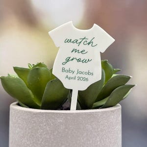 Może przedstawiać: Mała, jasnoszara betonowa doniczka z zieloną rośliną sukulentową. Biały znak w kształcie body niemowlęcego z napisem "watch me grow", imieniem "Baby Jacobs" i datą "April 2026".