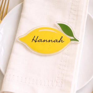Puede incluir: Una tarjeta de lugar con forma de limón amarillo con el nombre "Hannah" en escritura negra, colocada sobre una servilleta blanca encima de un plato blanco. Un tenedor dorado y un arreglo floral amarillo son visibles en el fondo. El mantel es a cuadros amarillos y blancos.