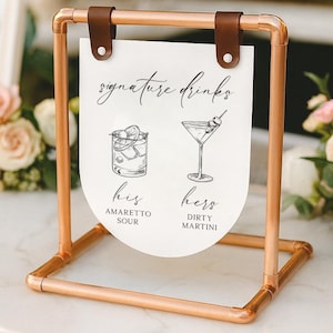 May include: A copper-colored metal sign holder with a white sign that reads "signature drinks." The sign features illustrations of an Amaretto Sour and a Dirty Martini. The sign is held in place by brown leather straps.