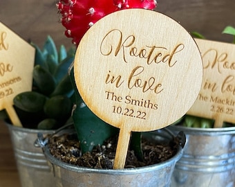 以下が含まれることがあります： 「Rooted in love」と書かれた木製の看板と植物が植えられた小さな金属製の鉢が3つあります。植物は多肉植物、サボテン、小さな緑色の植物です。看板には、カップルの名前と日付が書かれています。