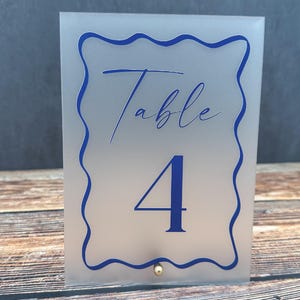 Puede incluir: Un número de mesa de acrílico esmerilado con un borde azul y el texto "Mesa 4" en una fuente azul de escritura.