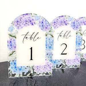 May include: Clear acrylic table number signs featuring a floral hydrangea design in purple and blue hues. Each sign displays the word "table" in script and a number. The signs have an arched top.