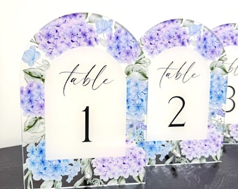 May include: Clear acrylic table number signs with a floral hydrangea design in shades of purple and blue. Each sign has the word "table" in script and a number. The signs are arched at the top.