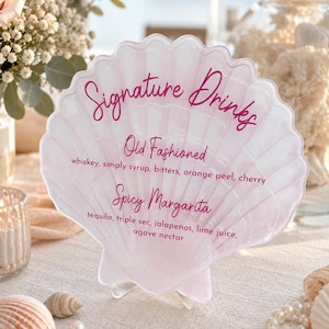May include: A translucent, light pink, shell-shaped acrylic sign featuring the text "Signature Drinks," "Old Fashioned" with ingredients, and "Spicy Margarita" with ingredients in dark pink script and print lettering, supported by a clear stand.