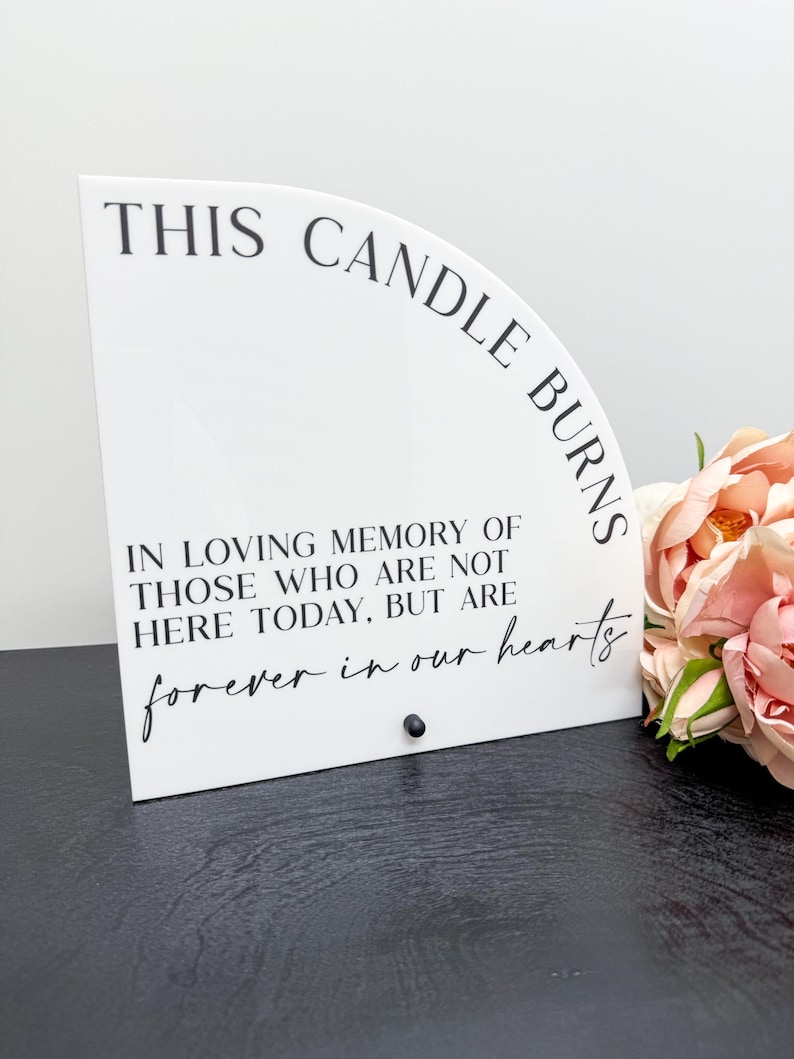 May include: A white, semi-circular memorial sign with the text "THIS CANDLE BURNS" and a heartfelt message. The sign includes a small black accent and is accompanied by pink and peach flowers.