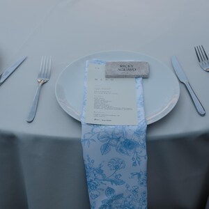 May include: A place setting with a white plate, silverware, and a blue and white floral napkin. A menu and a name card that reads "Ricky Aguayo" are on the plate. The table is covered with a white tablecloth.