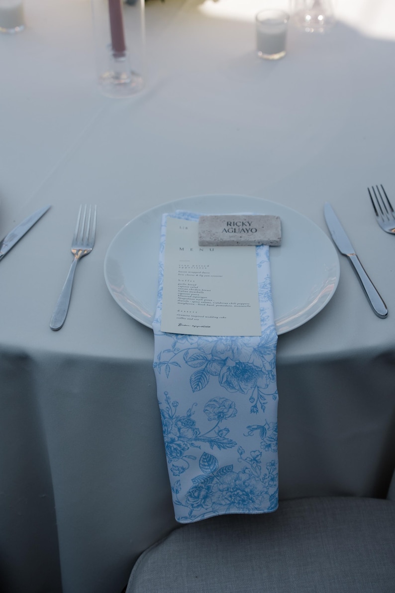 May include: A place setting with a white plate, silverware, and a blue and white floral napkin. A menu and a name card that reads "Ricky Aguayo" are on the plate. The table is covered with a white tablecloth.
