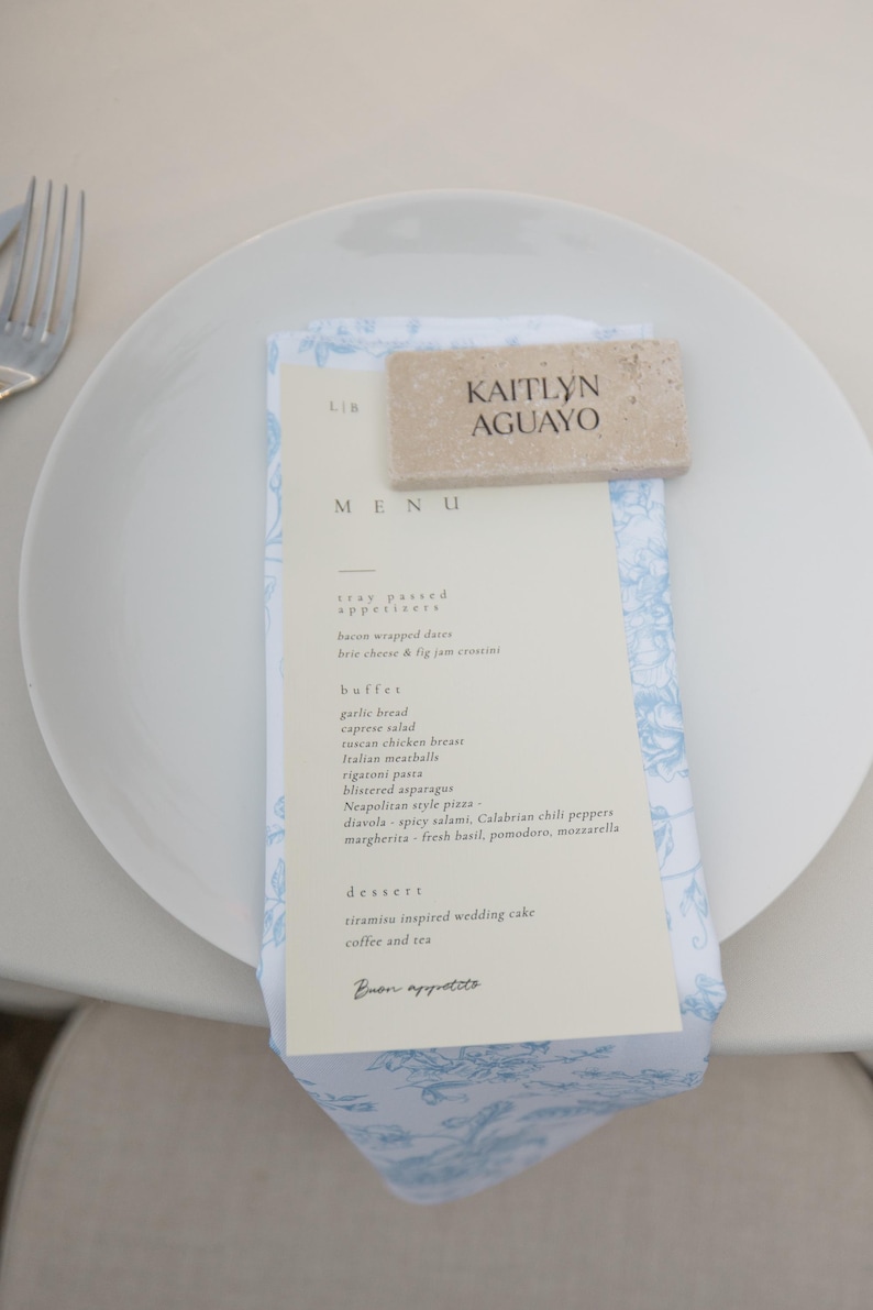 May include: A white plate with a menu and a napkin. The menu features a list of appetizers, buffet items, and desserts. A stone name card with the name "KAITLYN AGUAYO" rests on top. The napkin has a blue floral pattern.