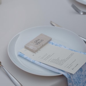 May include: A white plate with a menu and a napkin with a blue floral pattern. A small rectangular stone with the text "GINA WILLIG" is on top of the menu. Silverware is visible on the table.