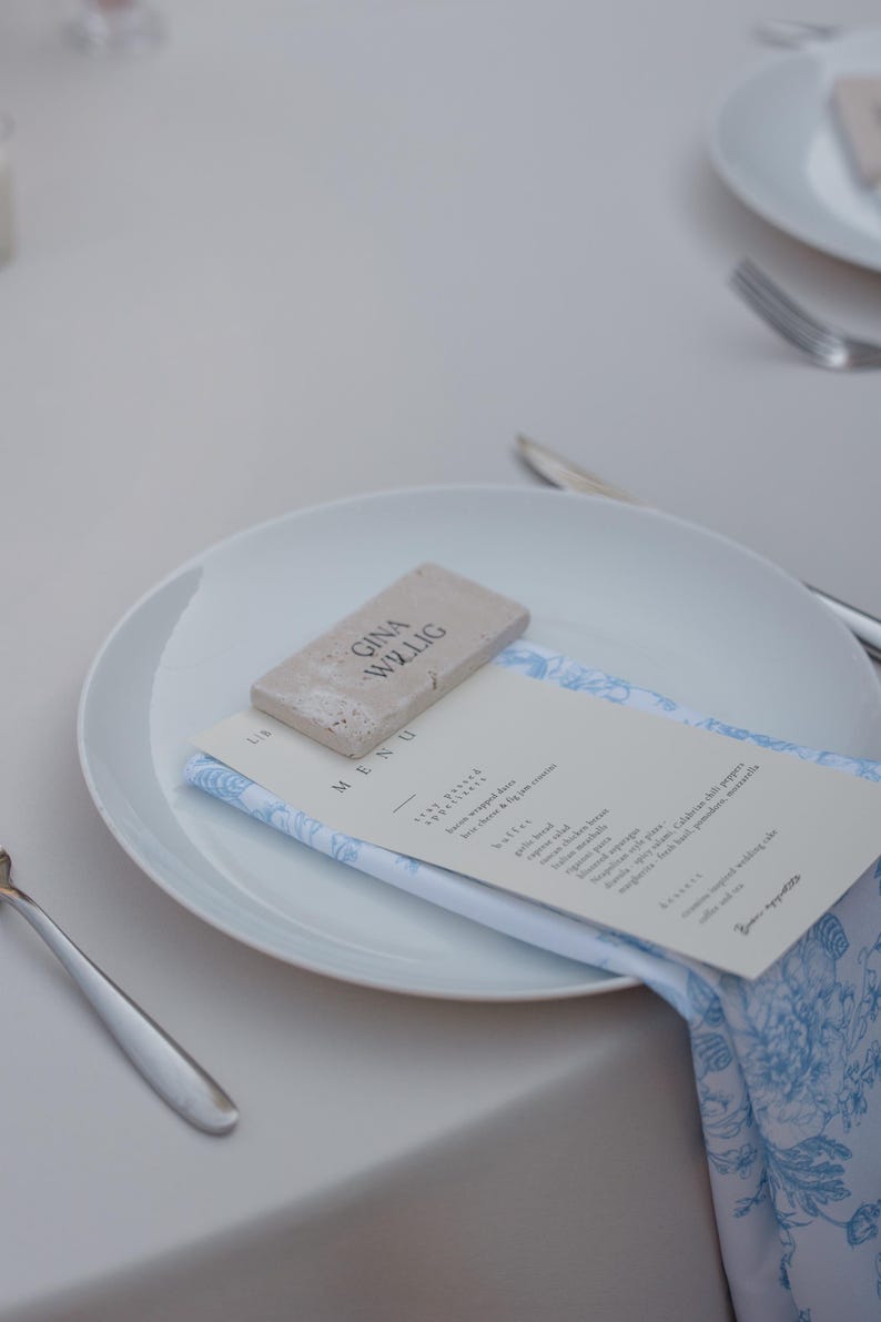 May include: A white plate with a menu and a napkin with a blue floral pattern. A small rectangular stone with the text "GINA WILLIG" is on top of the menu. Silverware is visible on the table.