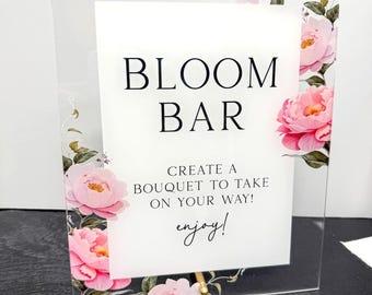 May include: Clear acrylic sign with the words "BLOOM BAR" in black, with the text "CREATE A BOUQUET TO TAKE ON YOUR WAY! enjoy!" below. The sign is decorated with pink floral accents.