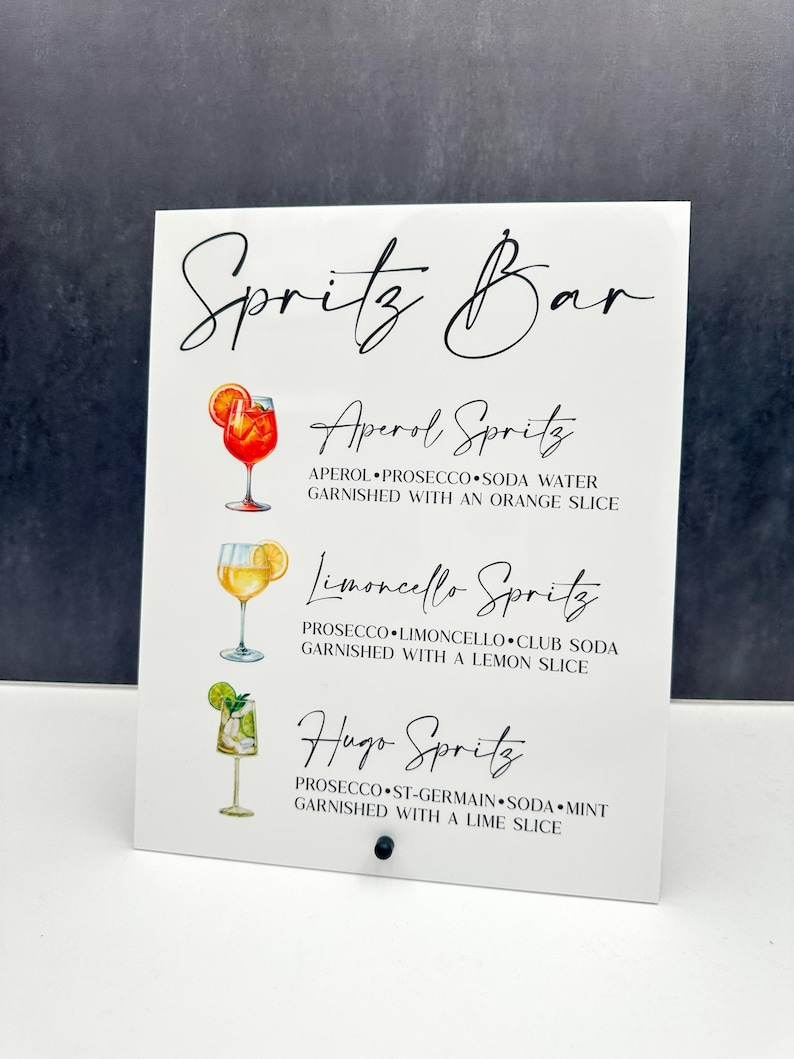 May include: White sign with black text listing three different spritz cocktails: Aperol Spritz, Limoncello Spritz, and Hugo Spritz. Each cocktail is illustrated with a watercolor drawing of a glass with the ingredients listed below.