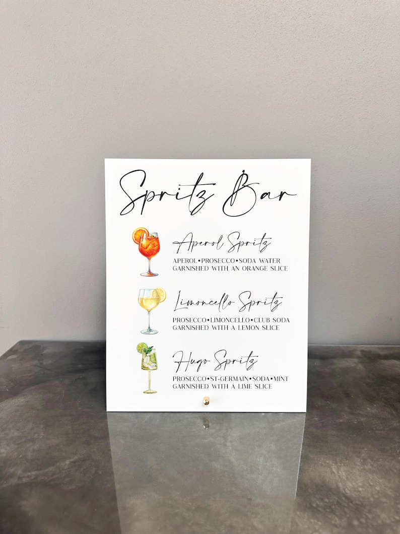 May include: A white sign with black text listing three different spritzes: Aperol Spritz, Limoncello Spritz, and Hugo Spritz. Each spritz has a small illustration of the drink next to it.