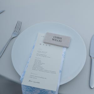 May include: A white plate with a menu and a napkin with a blue floral pattern. A small rectangular name card with the name "CHRIS WILLIG" is on top of the menu. Silverware is on either side of the plate.