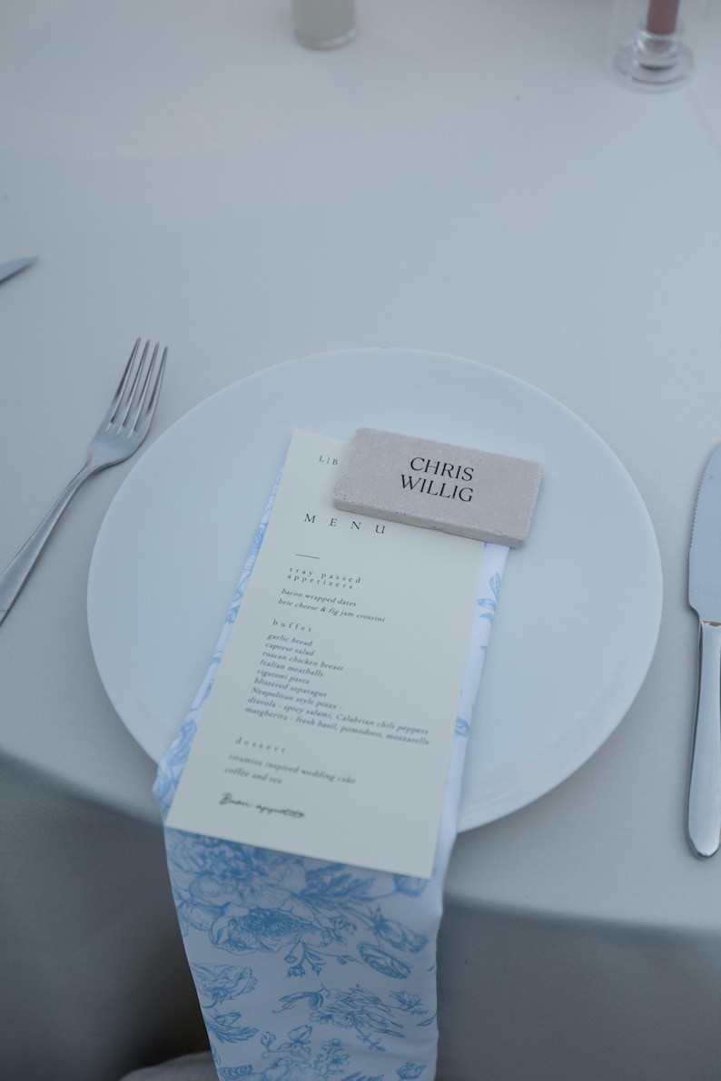 May include: A white plate with a menu and a napkin with a blue floral pattern. A small rectangular name card with the name "CHRIS WILLIG" is on top of the menu. Silverware is on either side of the plate.