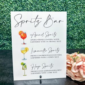 May include: A white sign with black text that reads "Spritz Bar" and lists three different spritz cocktails: Aperol Spritz, Limoncello Spritz, and Hugo Spritz. Each cocktail is illustrated with a glass of the drink.