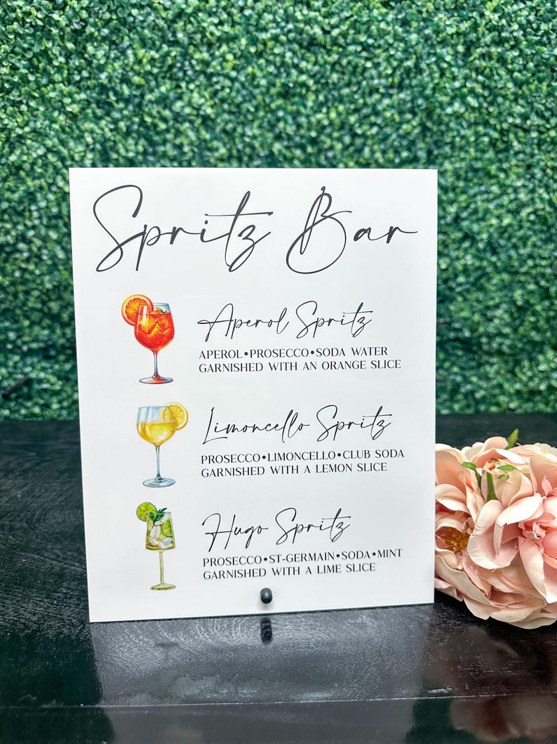 May include: A white sign with black text that reads "Spritz Bar" and lists three different spritz cocktails: Aperol Spritz, Limoncello Spritz, and Hugo Spritz. Each cocktail is illustrated with a glass of the drink.