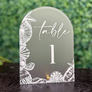 Puede incluir: Un número de mesa verde salvia con una parte superior arqueada. La palabra "table" está escrita en blanco sobre el número "1". Ilustraciones de conchas marinas blancas bordean los bordes del letrero. Un pequeño perno dorado está en la parte inferior.