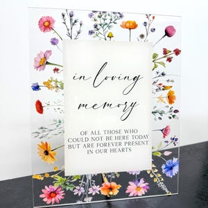 May include: A clear acrylic memorial sign with floral decoration. The sign displays the words "in loving memory" and a message about those who are forever present in our hearts. The edges are adorned with colourful flowers in pink, orange, and blue.