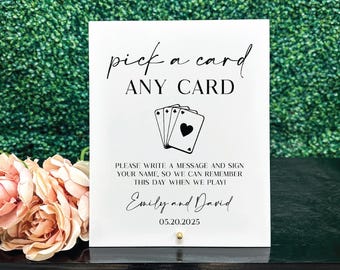 May include: White sign with black text that reads "pick a card ANY CARD".  The sign also features a black and white illustration of playing cards.  The text below the illustration reads "PLEASE WRITE A MESSAGE AND SIGN YOUR NAME, SO WE CAN REMEMBER THIS DAY WHEN WE PLAY! Emily and David 05.20.2025"