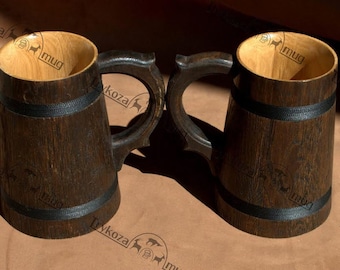 Könnte beinhalten: Zwei Bierkrüge aus Holz mit schwarzen Bändern um die Mitte. Die Krüge sind aus dunklem Holz gefertigt und haben eine natürliche Holzoptik. Die Krüge stehen auf einer braunen Oberfläche.