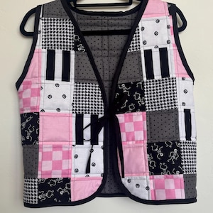 May include: A quilted vest with a patchwork design. The vest features squares of pink, black, white, and gray fabrics with various patterns, including houndstooth, stripes, and skeleton motifs. It has a black trim and a tie closure.