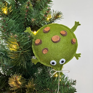 May include: A whimsical, handmade Christmas tree ornament shaped like a green, spotted alien. The felt alien has brown spots, white eyes, and small arms and legs. It is attached to a Christmas tree with a string.