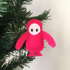 May include: A handmade, red felt ornament in the shape of a character from the video game Fall Guys, hanging on a Christmas tree. The ornament has a white face with two black eyes and a stitched outline.
