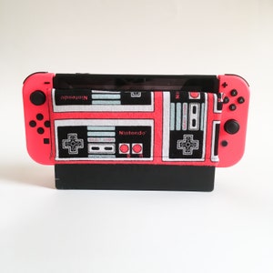 May include: A pink Nintendo Switch console with a black screen and a fabric cover featuring a retro controller design. The cover has a black background with white and light blue controller graphics and the word "Nintendo".