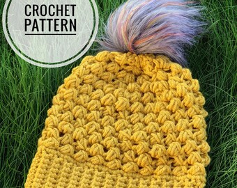 May include: A crocheted beanie hat in a mustard yellow color with a fluffy purple and white pom pom on top. The hat is lying on a bed of green grass. The text "CROCHET PATTERN" is visible in a white circle in the top left corner of the image.