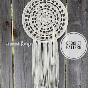 May include: A white crochet dreamcatcher with a circular design and a long fringe. The dreamcatcher is hanging on a wooden fence. The text "CROCHET PATTERN" is visible in a circle on the right side of the image.