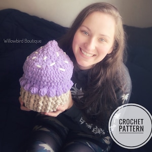 Può includere: Una donna tiene un cupcake all'uncinetto viola e bianco con codette bianche. Il cupcake è un giocattolo di peluche. Il testo "CROCHET PATTERN" è in un cerchio nell'angolo in basso a destra dell'immagine. Il testo "Willowbird Boutique" è nell'angolo in alto a sinistra dell'immagine.