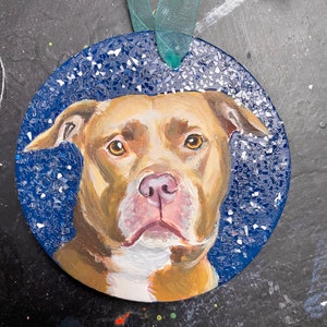 May include: A round, blue and white ornament with a painted image of a brown and white pit bull dog. The ornament has a green ribbon attached.