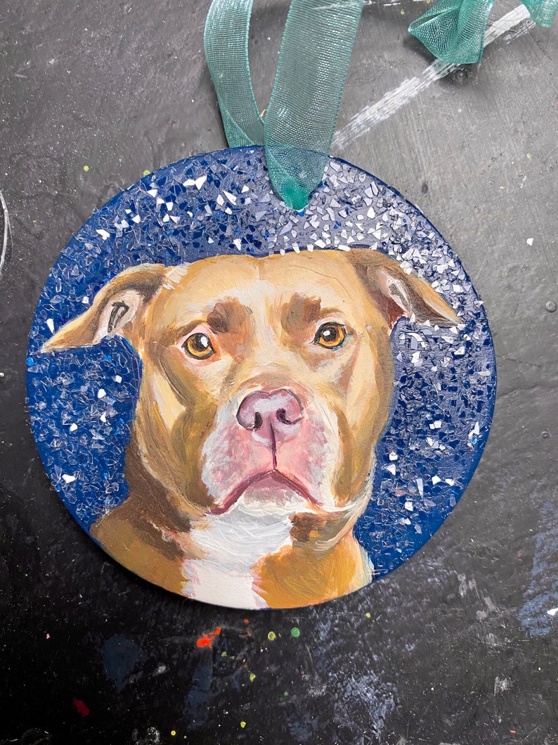 May include: A round, blue and white ornament with a painted image of a brown and white pit bull dog. The ornament has a green ribbon attached.