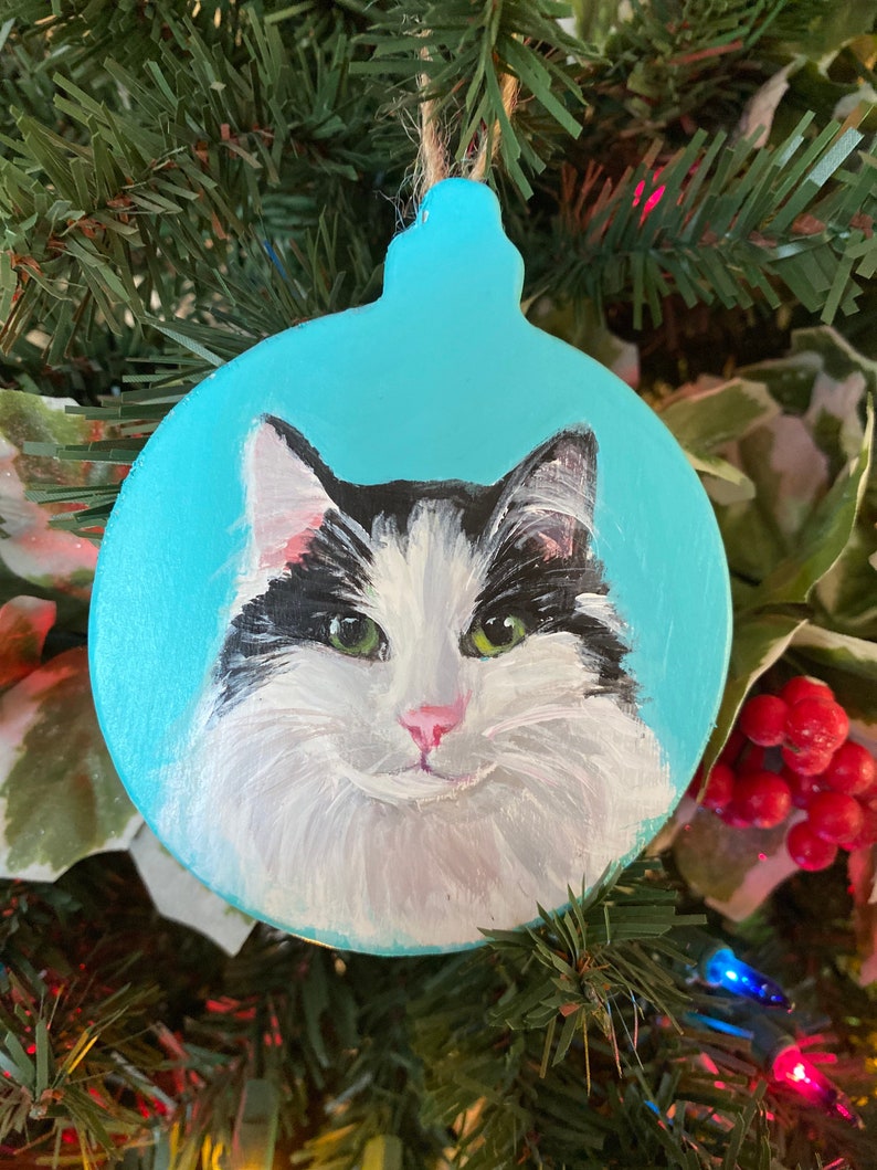 May include: A hand-painted wooden Christmas ornament featuring a black and white cat with green eyes. The ornament is painted on a light blue background.