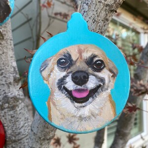 May include: A hand-painted Christmas ornament featuring a dog's face on a blue background. The dog is smiling with its tongue sticking out.