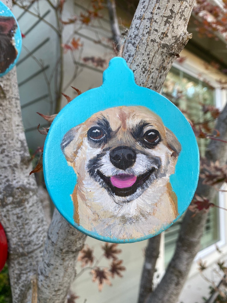 May include: A hand-painted Christmas ornament featuring a dog's face on a blue background. The dog is smiling with its tongue sticking out.