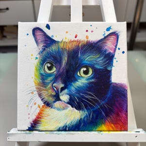 May include: A colorful painting of a cat's face on a white canvas. The cat has black fur with white markings and green eyes. The background is white with colorful paint splatters.